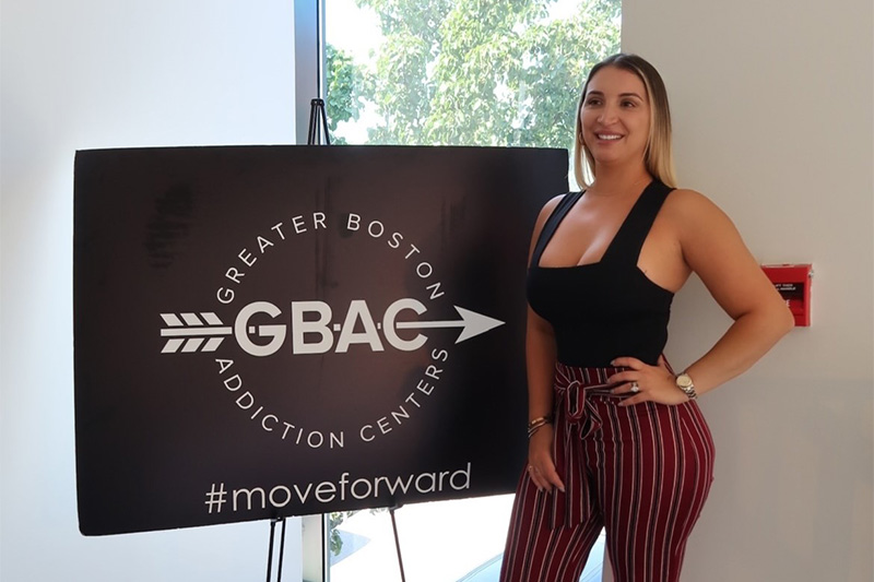 A woman stands proudly in front of Greater Boston Addiction Centers sign. The sign reads: "GBAC - Greater Boston Addiction Center #moveforward"